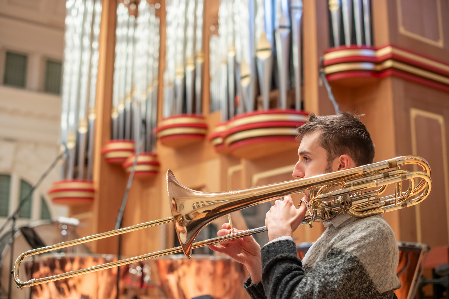 A male student performing the trombone in an orchestra rehearsal, with an organ behind them.